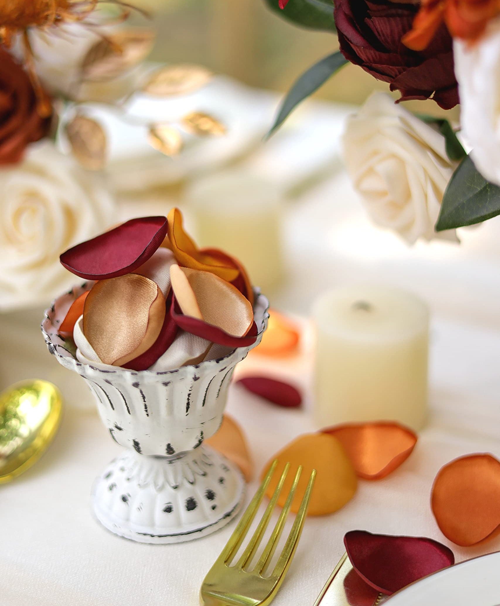 Terracotta satin rose petals in a decorative white vase with gold fork and candles on a table