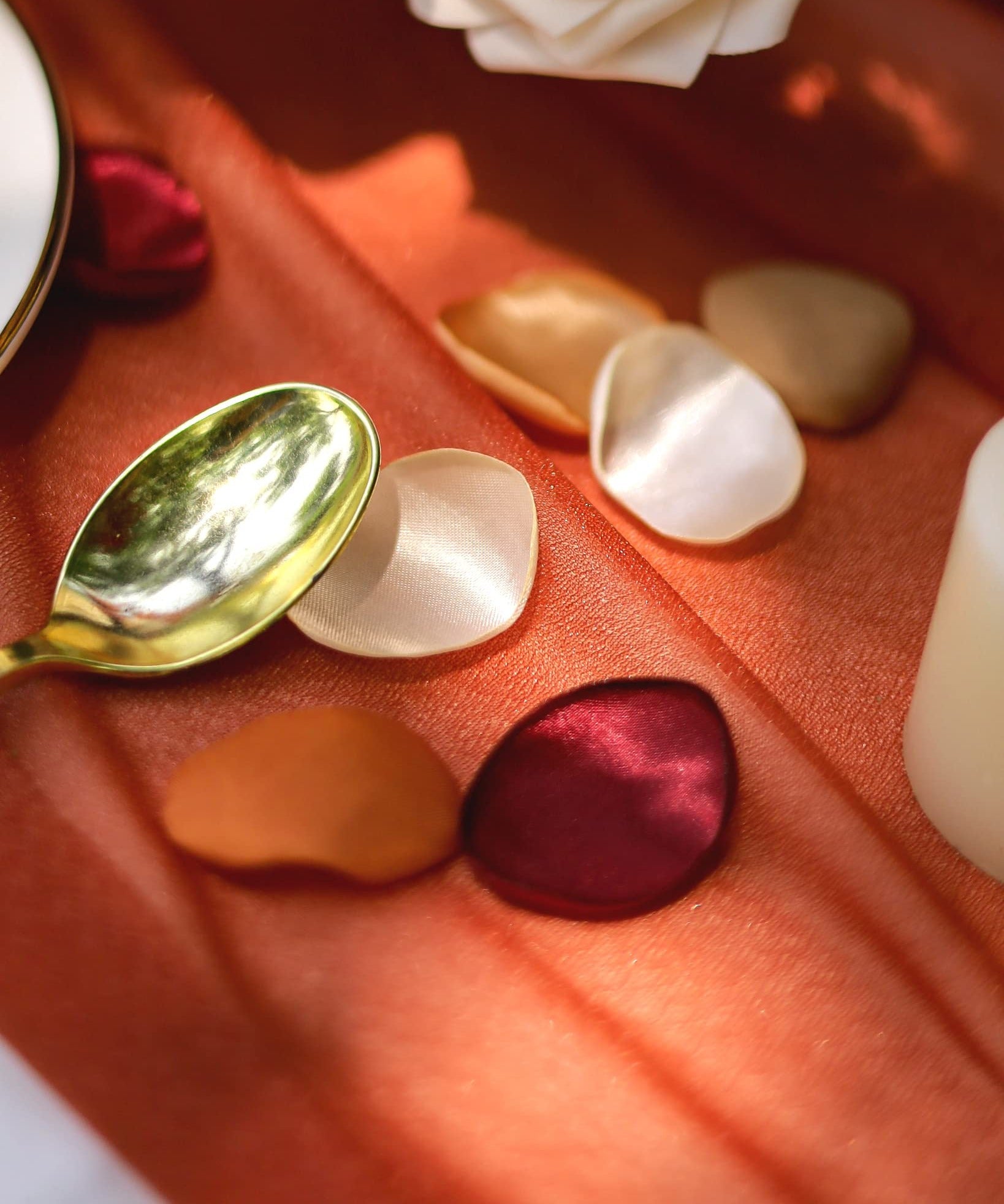 Terracotta satin rose petals scattered on an orange tablecloth with a gold spoon and white roses