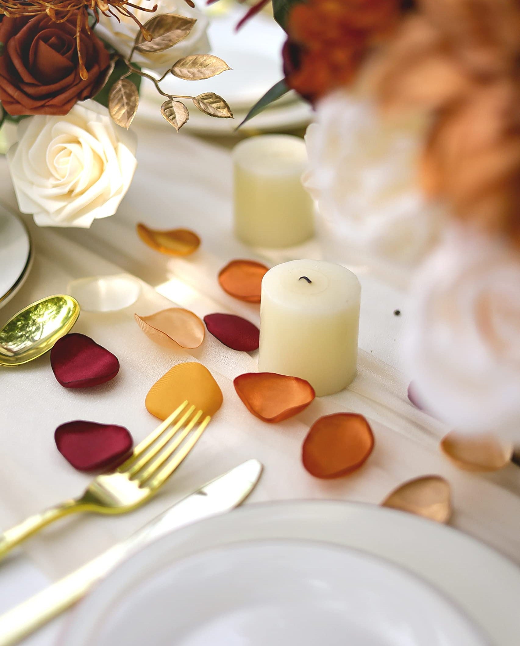 Terracotta satin rose petals scattered on a table with candles and gold cutlery, arranged for a wedding.