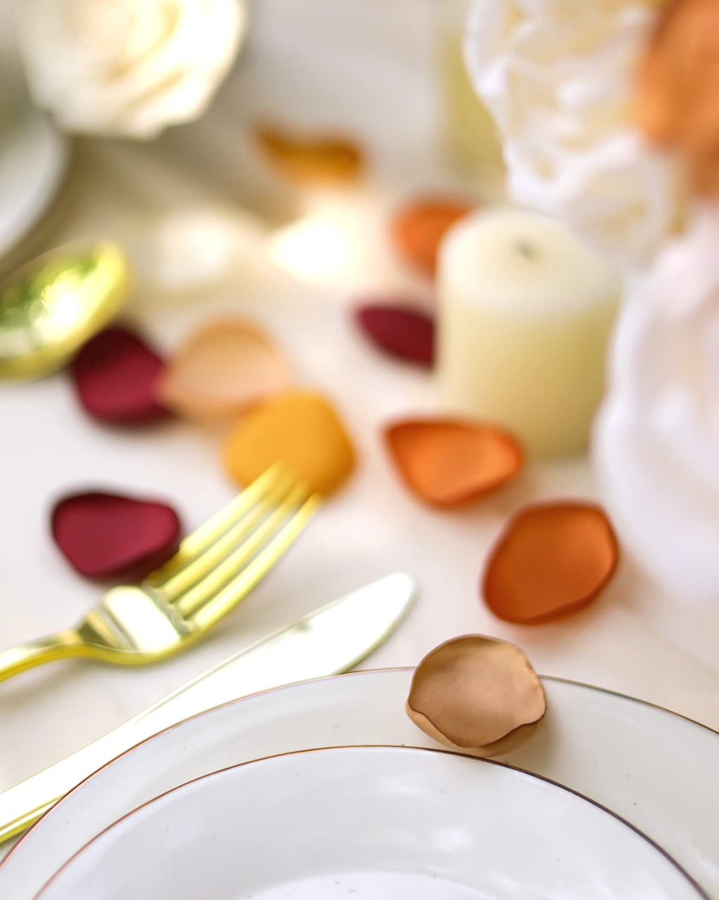 Terracotta satin rose petals scattered on a table setting with plates, gold cutlery, and a candle.