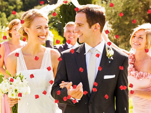 Bride and groom smiling as red rose petals fall around them during an outdoor wedding ceremony.