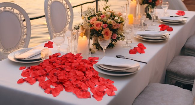 Dark red artificial rose petals scattered on a white tablecloth set for a romantic dinner with candles and flowers.