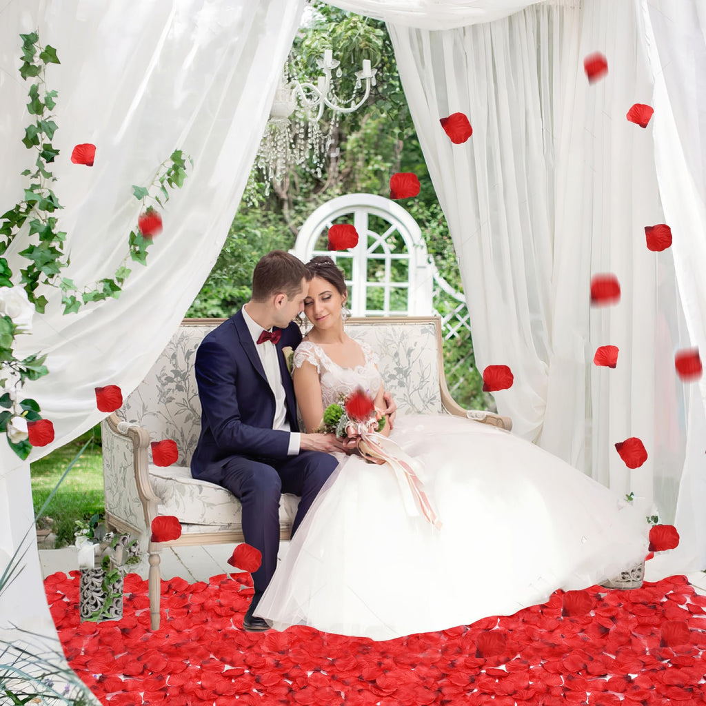 Bride and groom sitting on a decorative couch surrounded by red rose petals, with petals falling from above.