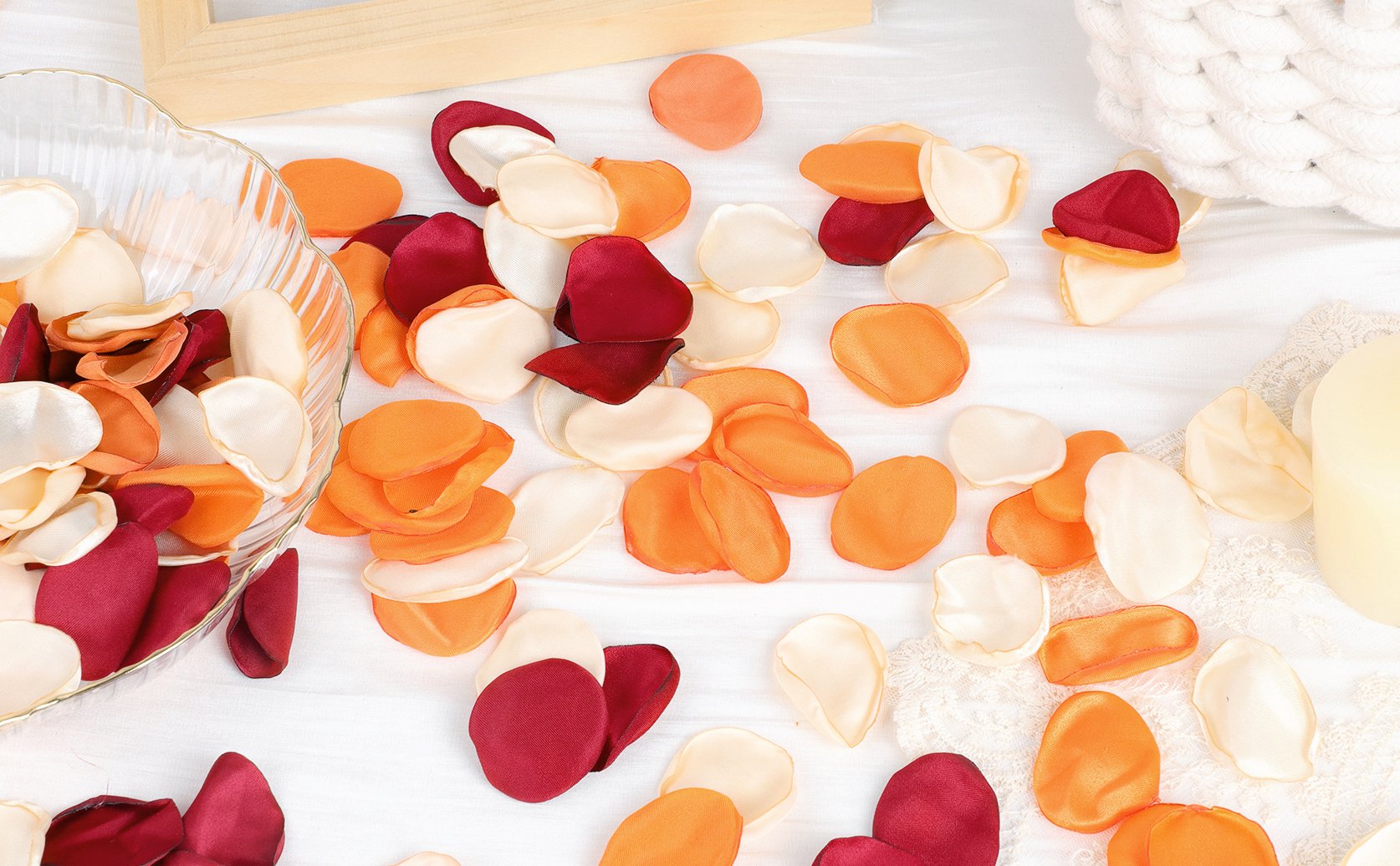 Assorted orange, cream, and burgundy artificial rose petals scattered on a white surface with a glass bowl.