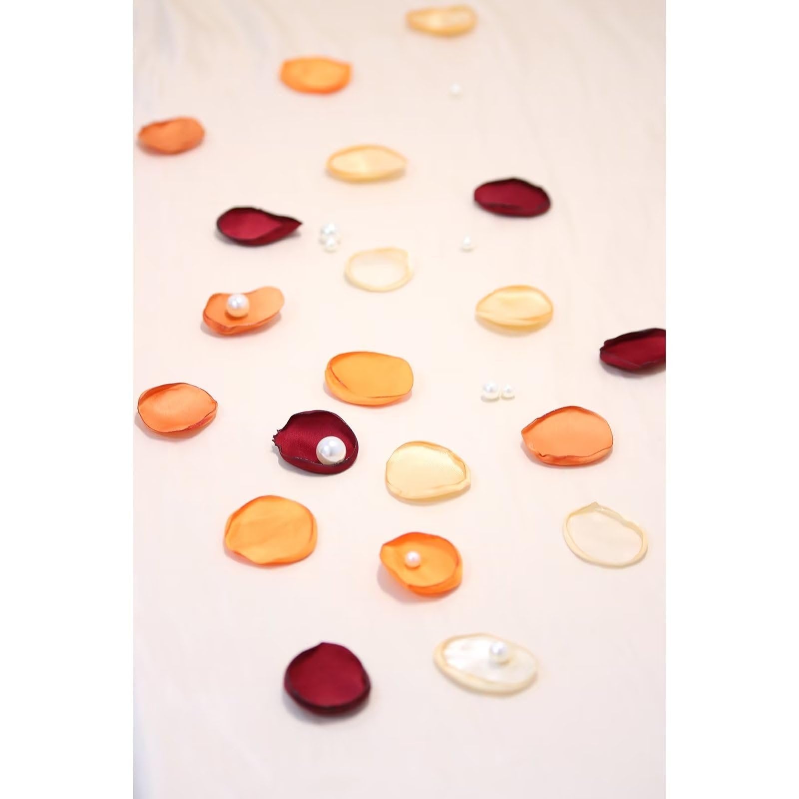 Assorted orange, cream, and burgundy artificial rose petals scattered with small pearls on a light fabric surface.