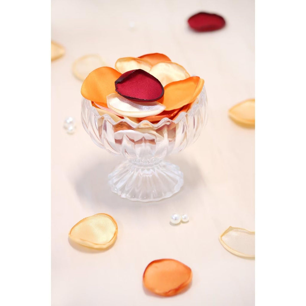 Assorted orange, cream, and burgundy artificial rose petals in a clear glass dish with scattered petals and pearls on a light background.