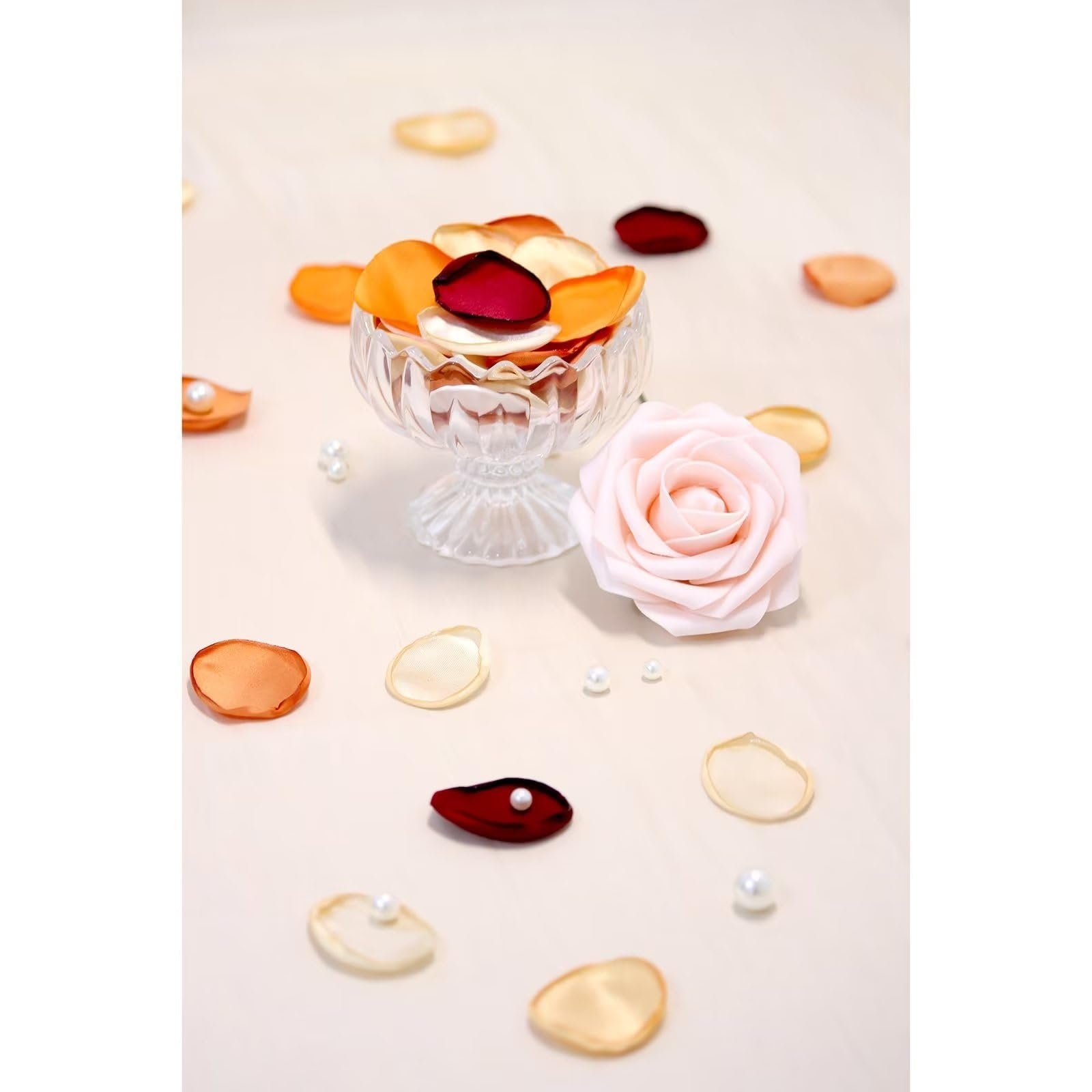 Assorted orange, cream, and red artificial rose petals scattered around a glass dish and a pink rose.
