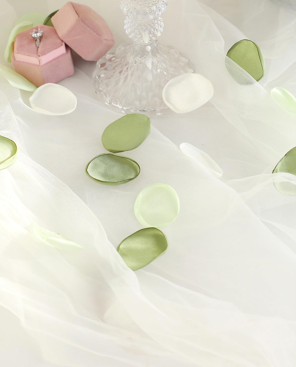 Faux silk rose petals in pale green and white scattered on white tulle with a ring in a pink box and a candle in a crystal holder.