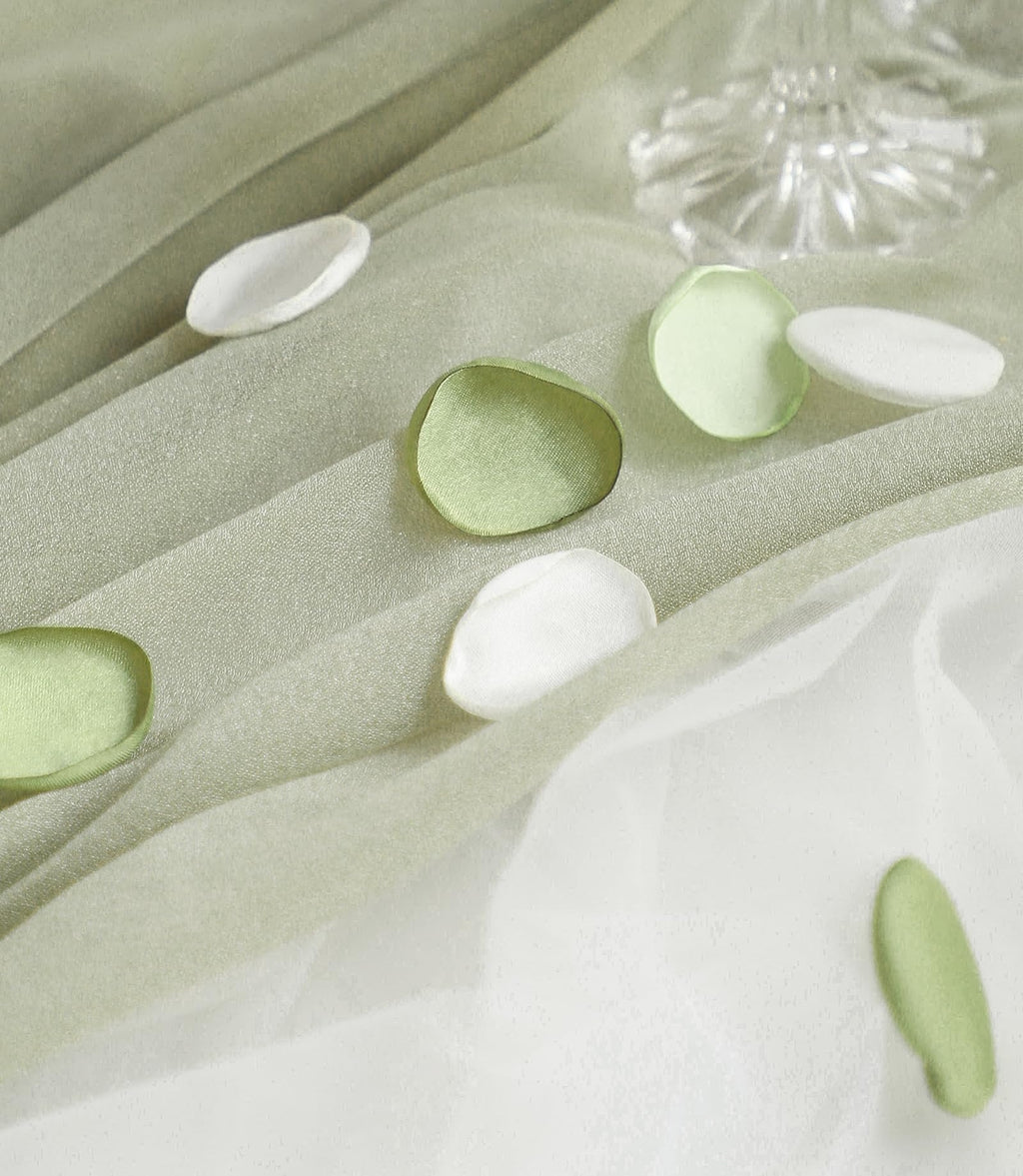 Faux silk rose petals in pale green and white scattered on draped fabric with a crystal vase in the background.