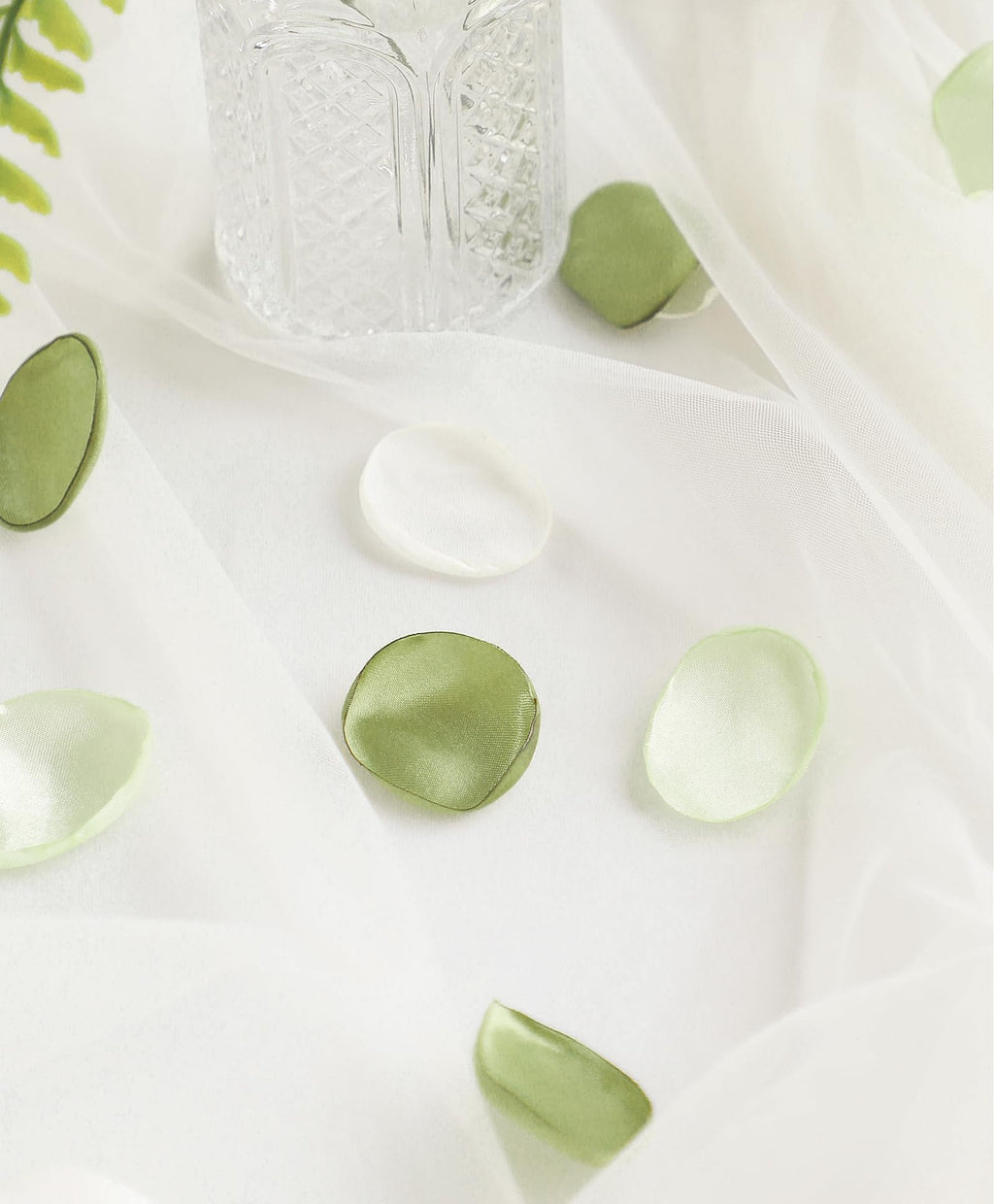 Faux silk rose petals in pale green and white scattered on sheer fabric with a crystal vase and greenery
