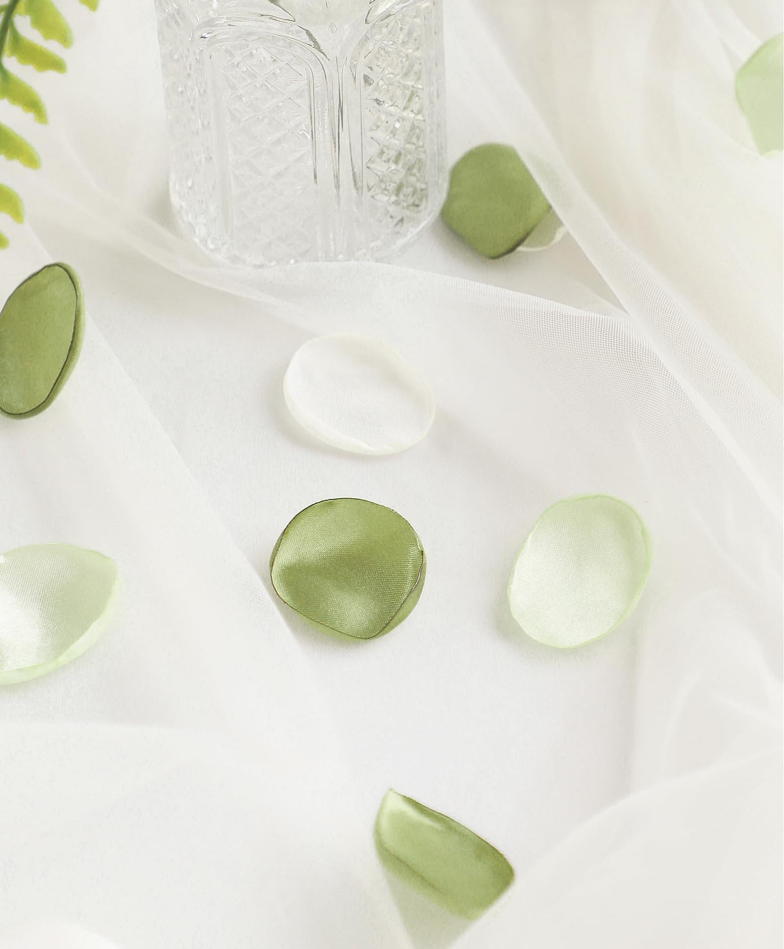 Faux silk rose petals in pale green and white scattered on sheer fabric with a crystal vase and greenery