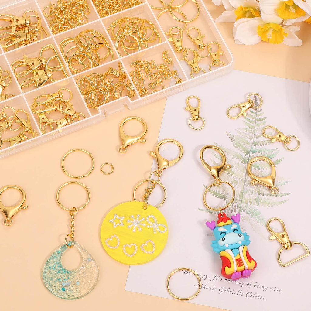 Gold keychain making kit components including clips, rings, chains, and charms, laid out with a decorative paper and flowers.