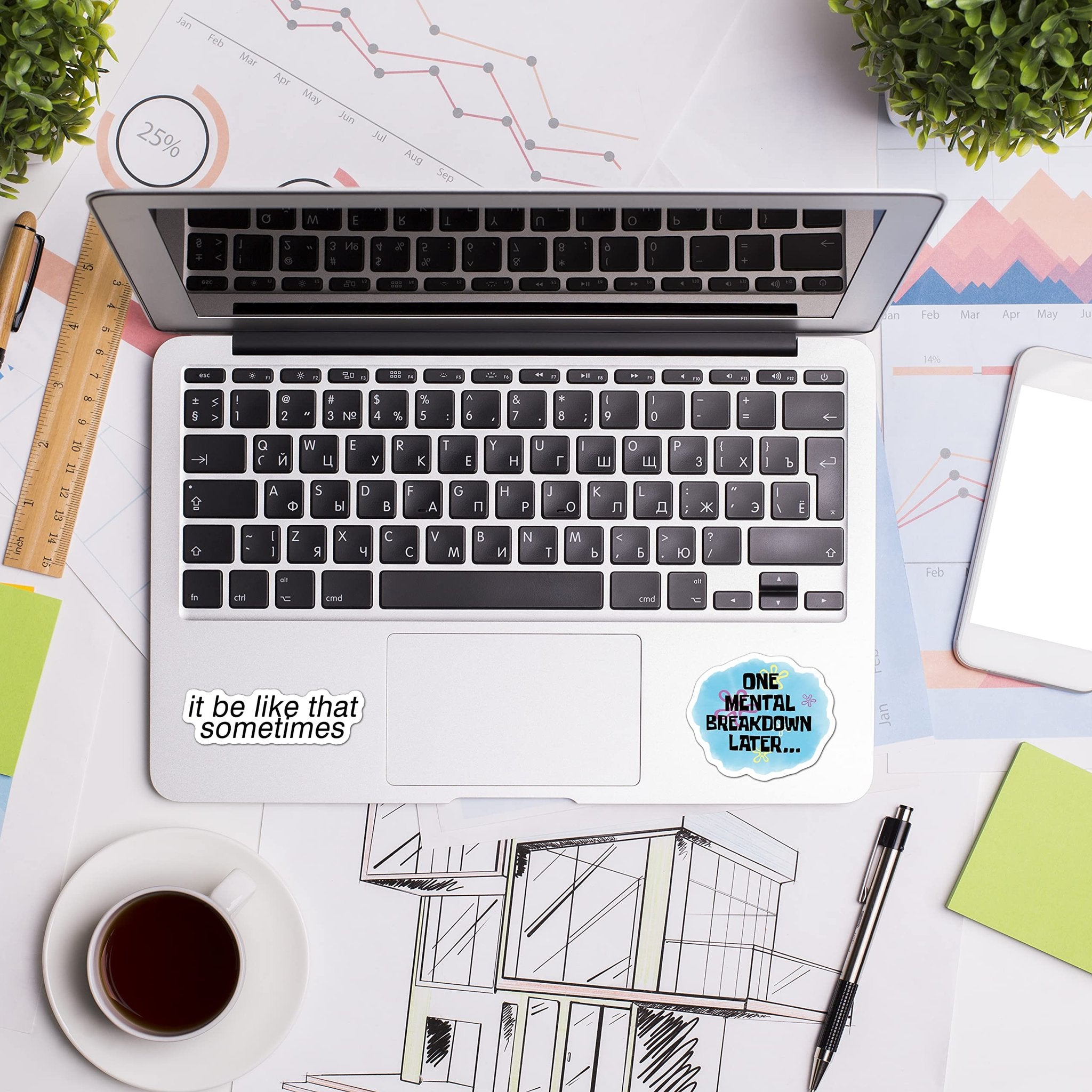 Two meme stickers on a laptop keyboard: 'it be like that sometimes' and 'ONE MENTAL BREAKDOWN LATER...'. The laptop is open on a desk with charts, a cup of coffee, and a drawing.