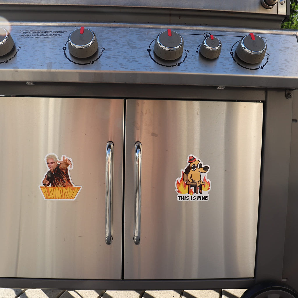 Two meme stickers applied to the stainless steel doors of a gas grill. One sticker depicts Guy Fieri with "FLAVORTOWN" text, and the other shows a dog in a burning room with "THIS IS FINE" text.