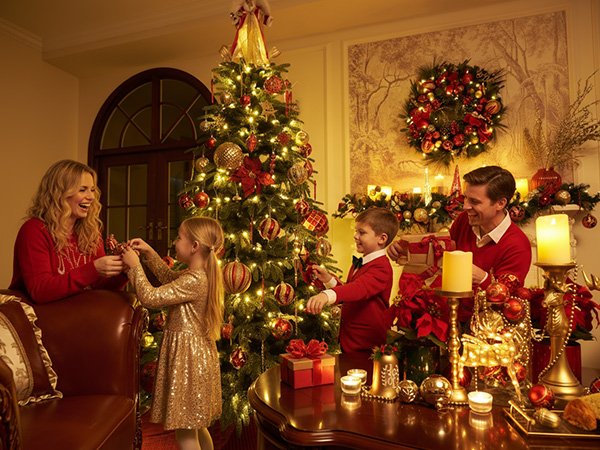 Family decorating a Christmas tree with red and gold ornaments in a festive living room setting.