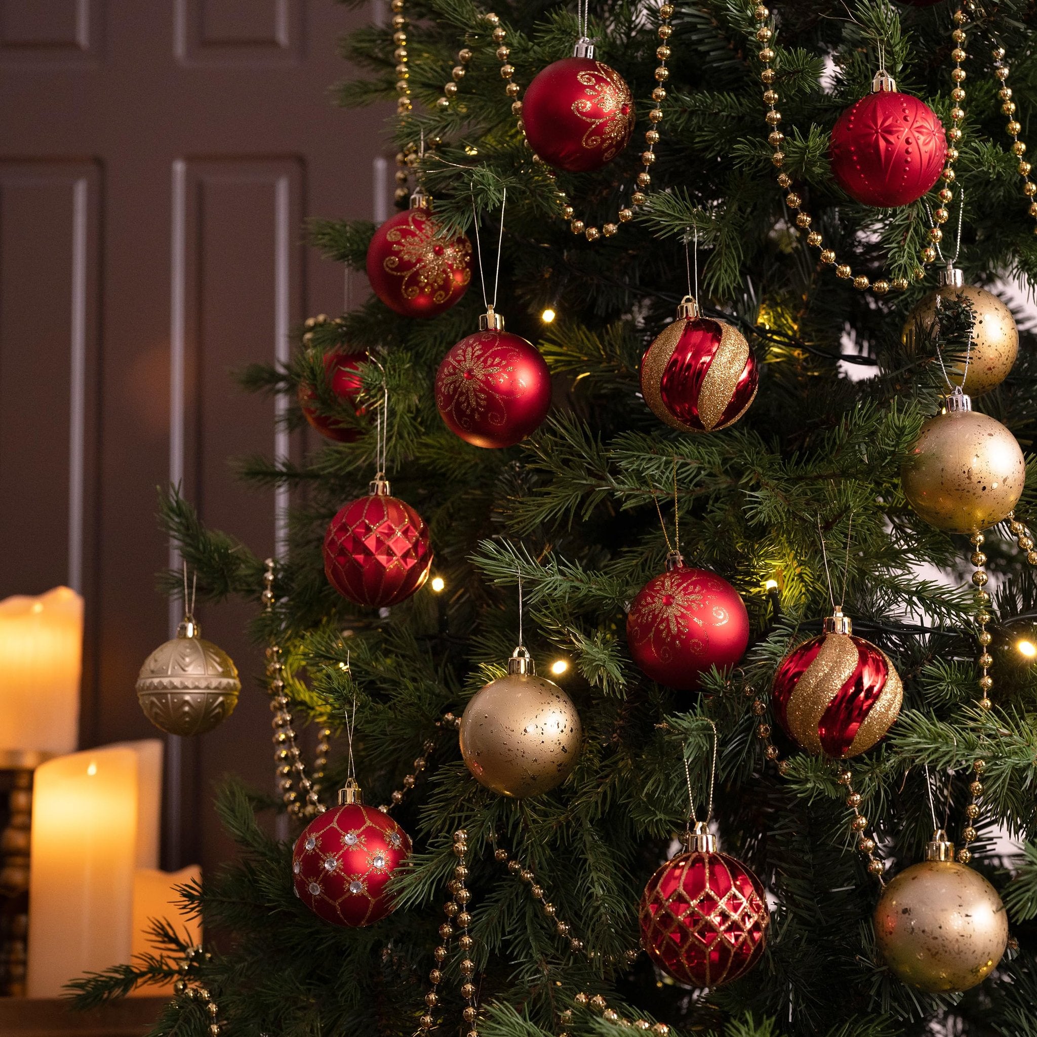 Red and gold shatterproof Christmas ball ornaments hanging on a decorated tree with candles in the background.