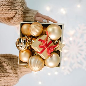 Person in sweater holding a box of gold shatterproof Christmas ball ornaments and a small gift box.