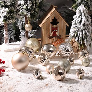 Assortment of shatterproof plastic Christmas ball ornaments in various sizes and finishes, arranged on a snowy surface with miniature trees and a wooden birdhouse.
