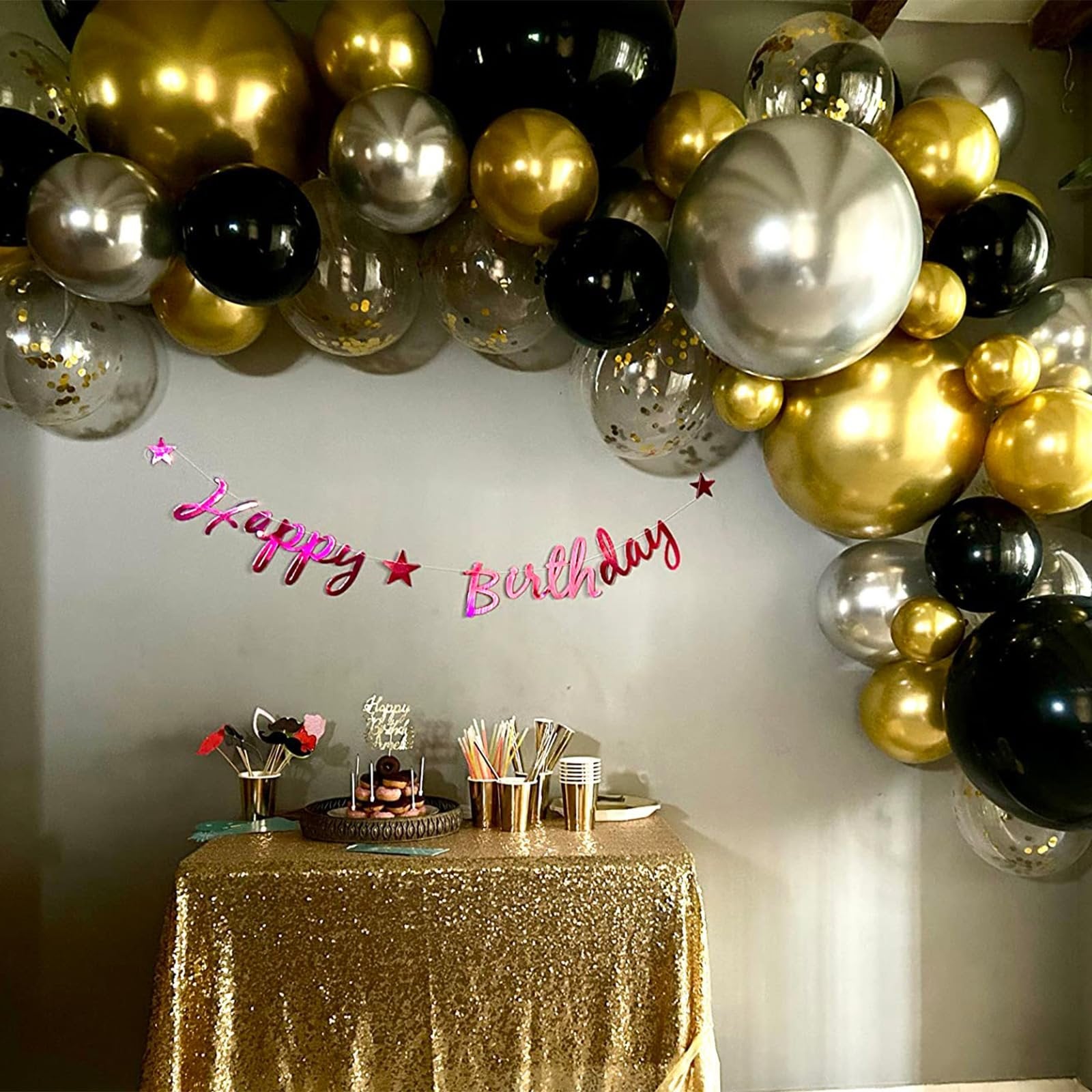 Birthday party setup with black, gold, and silver balloons arching over a table with a cake and party supplies.
