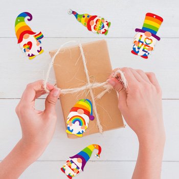 Hands tying a gift box with twine, surrounded by five rainbow gnome pride ornaments on a white wooden surface.