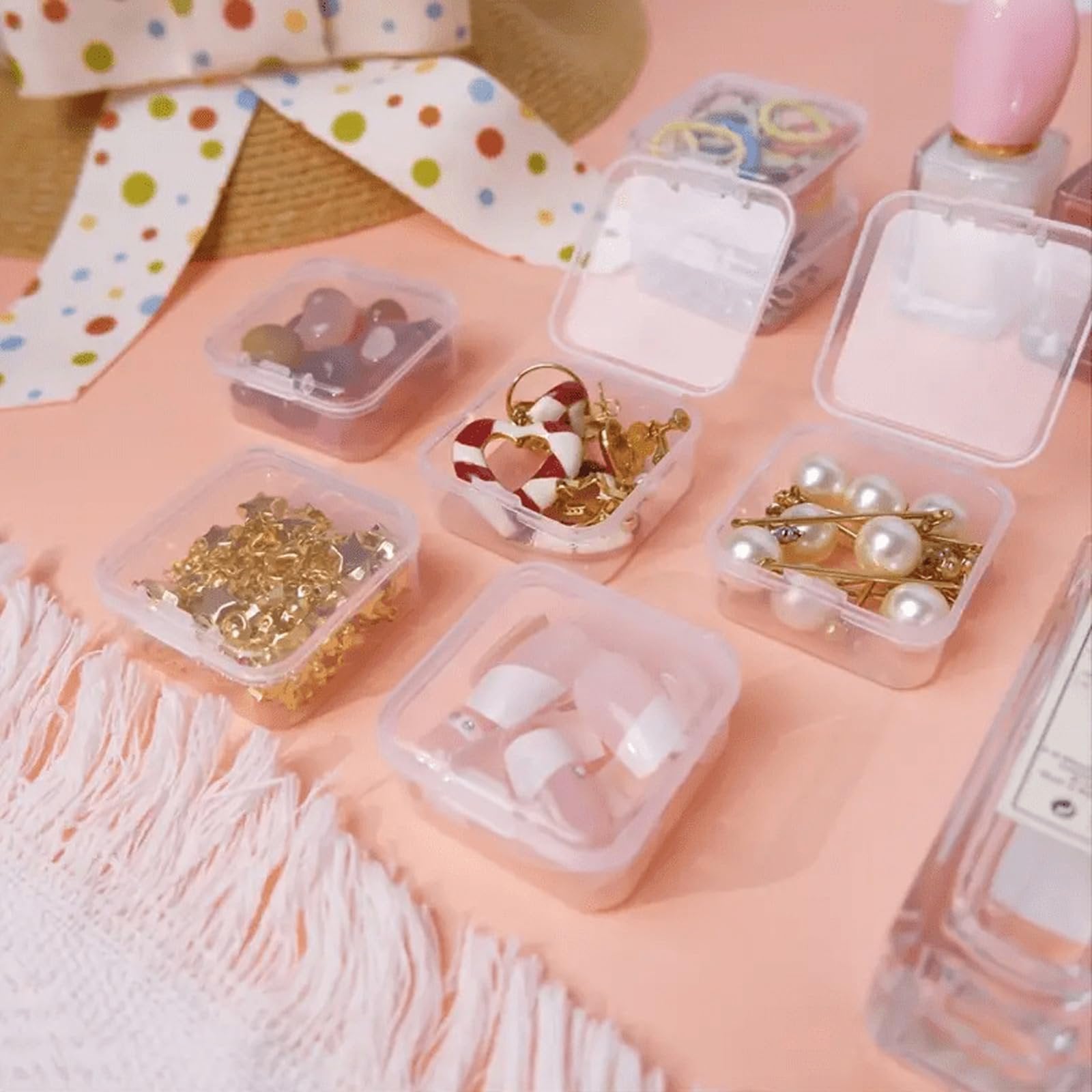 Clear plastic organizer boxes filled with beads, jewelry, and artificial nails on a pink surface with a decorative ribbon.