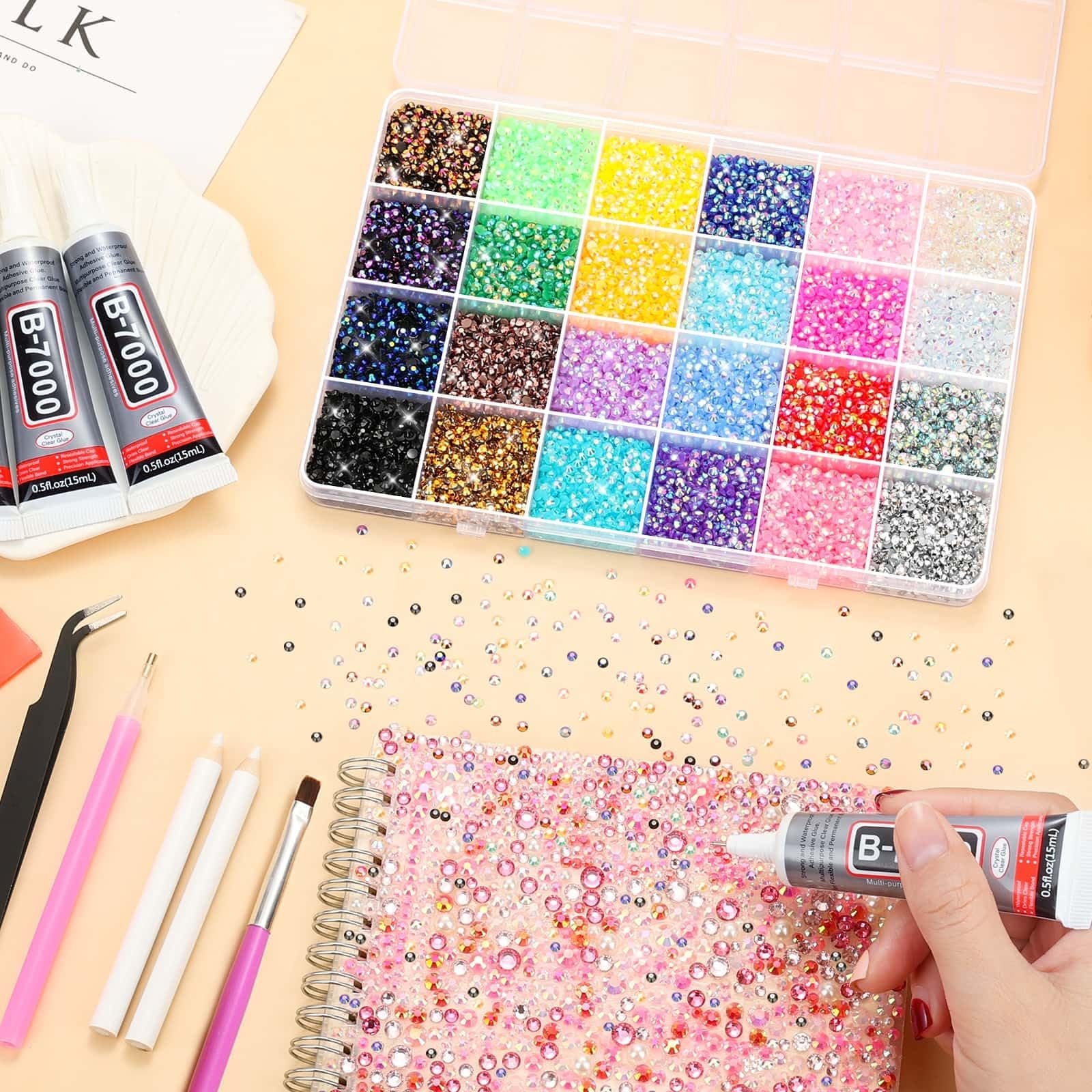 Flat lay of a DIY craft kit including a case of colorful rhinestones, two tubes of B-7000 adhesive glue, tweezers, and a notebook decorated with rhinestones.