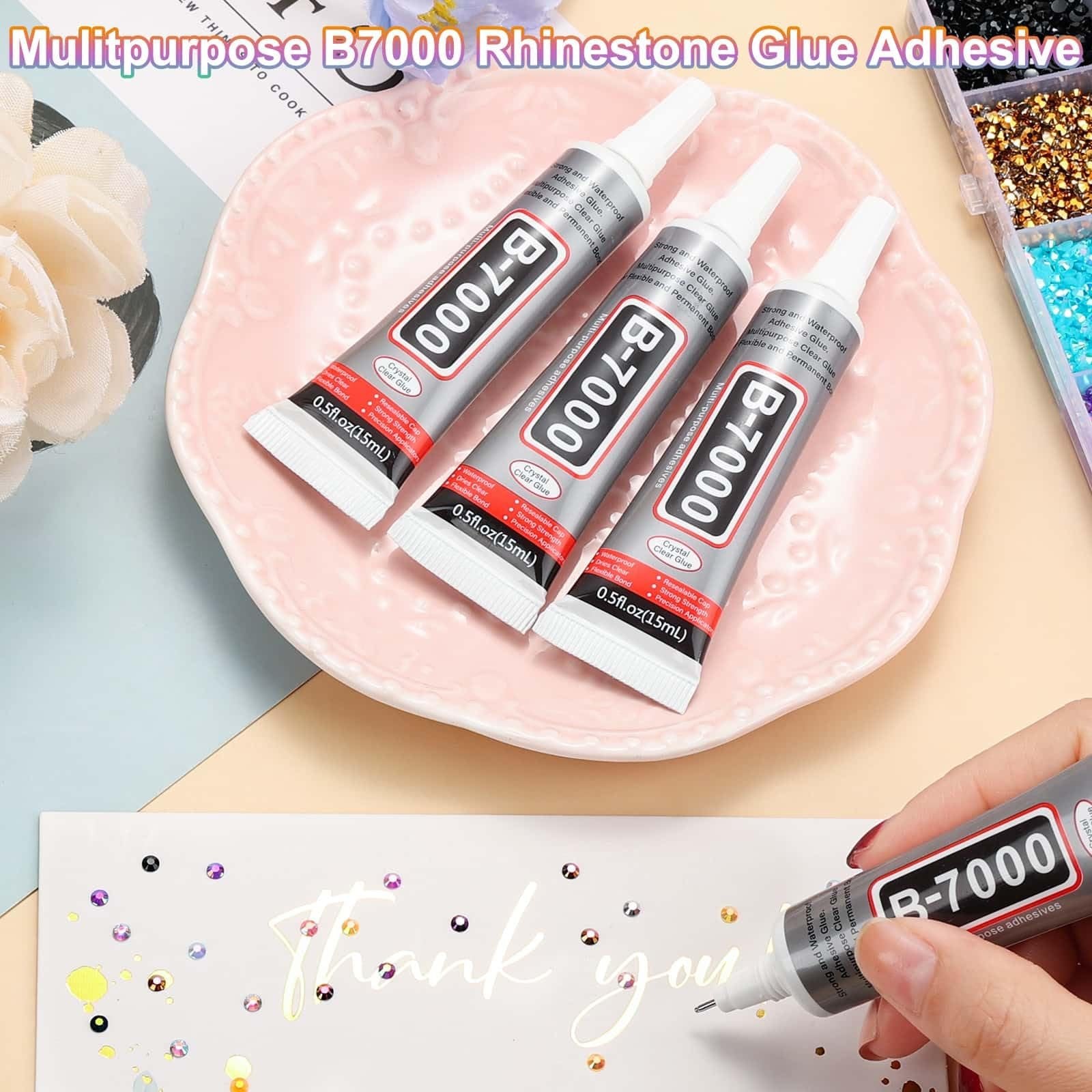 Three tubes of B-7000 clear adhesive glue on a pink plate, with a hand holding another tube over a 'Thank You' card with rhinestones.