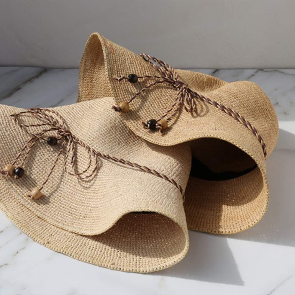 Two woven straw sun hats with decorative brown braided trim and wooden beads, displayed on a marble surface.