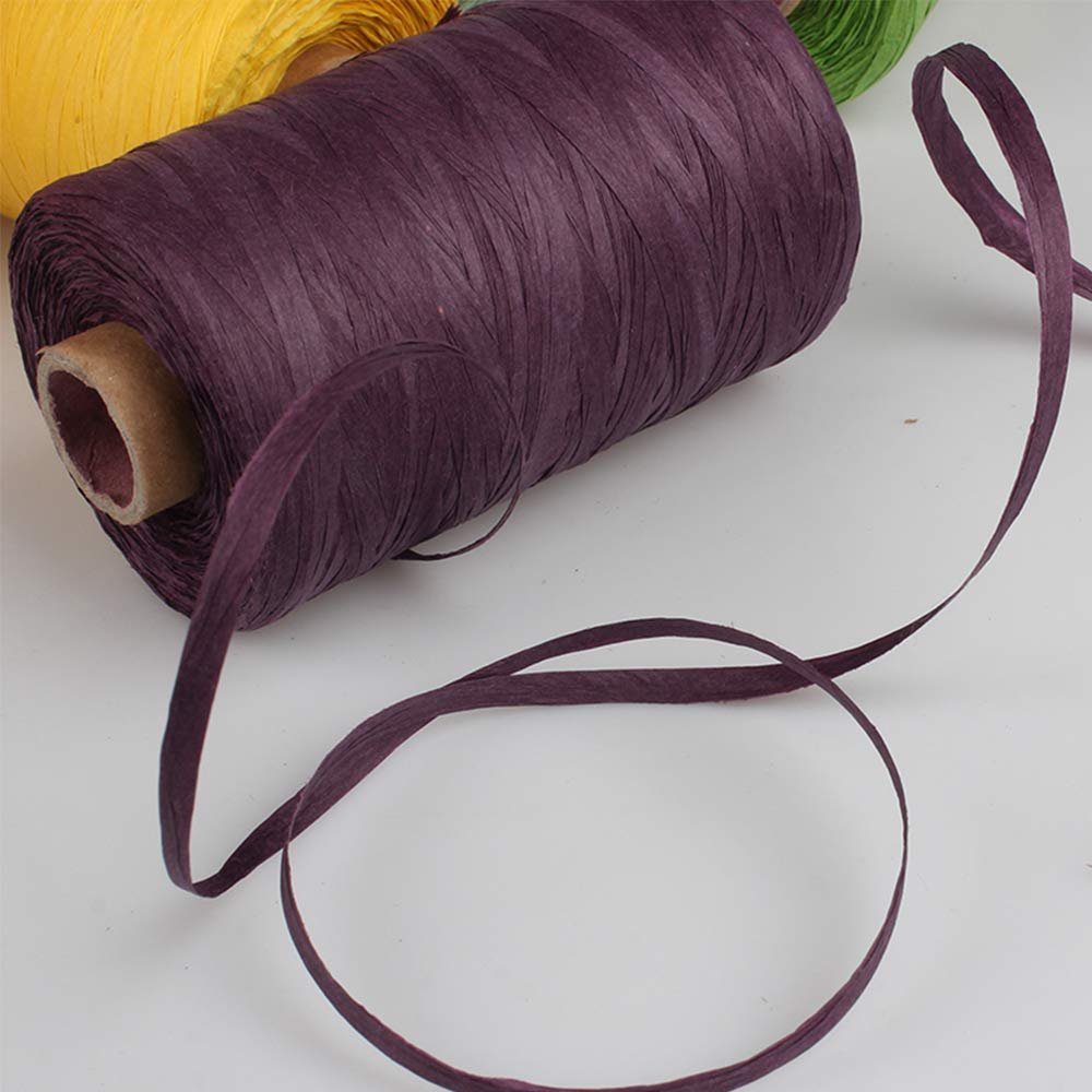 Spool of dark purple raffia paper yarn with a strand unspooling, other spools in yellow and green visible in background.
