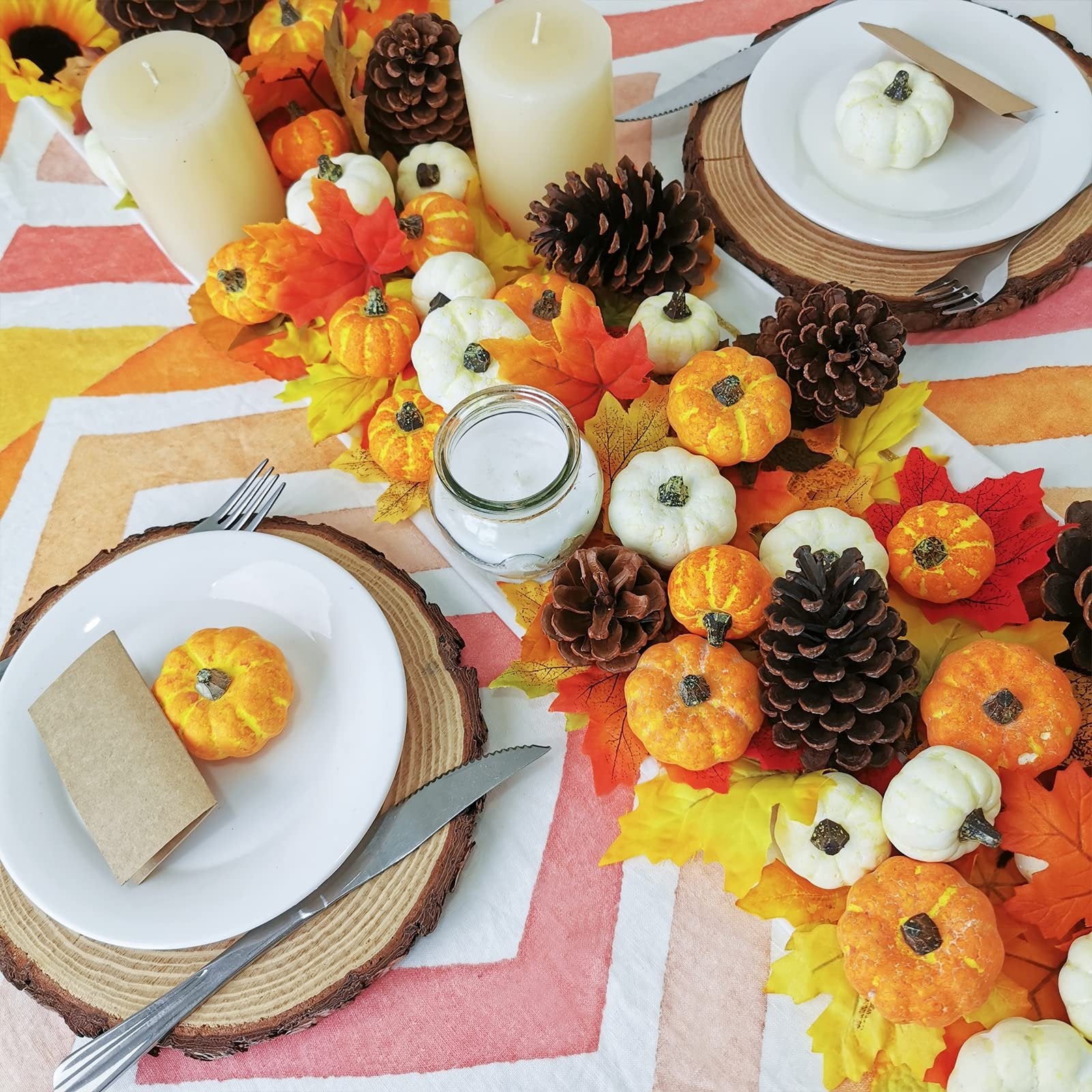 Assorted orange and cream mini pumpkins and pinecones arranged on a table runner with autumn leaves, set for a meal.