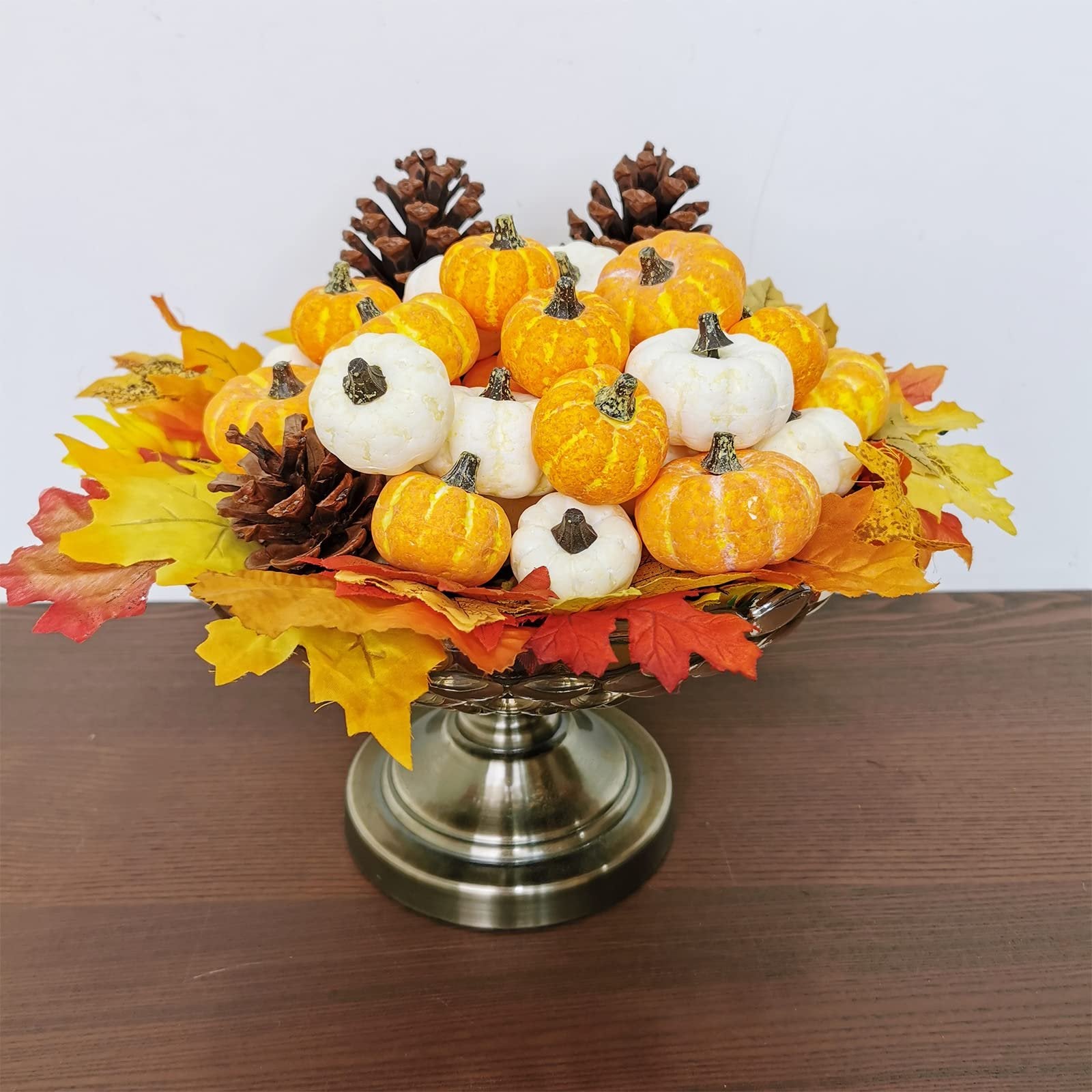 Assorted orange and cream mini pumpkins with pine cones and fall leaves in a decorative silver pedestal bowl.