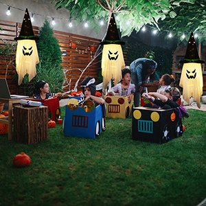 Four glowing witch hat ghost decorations hanging from trees at night with children in costume in a backyard.