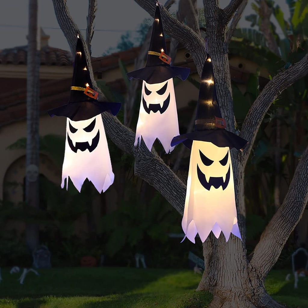 Three glowing ghost witch hat Halloween decorations hanging from a tree branch at dusk.