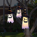 Three glowing ghost witch hat Halloween decorations hanging from a tree branch at dusk.