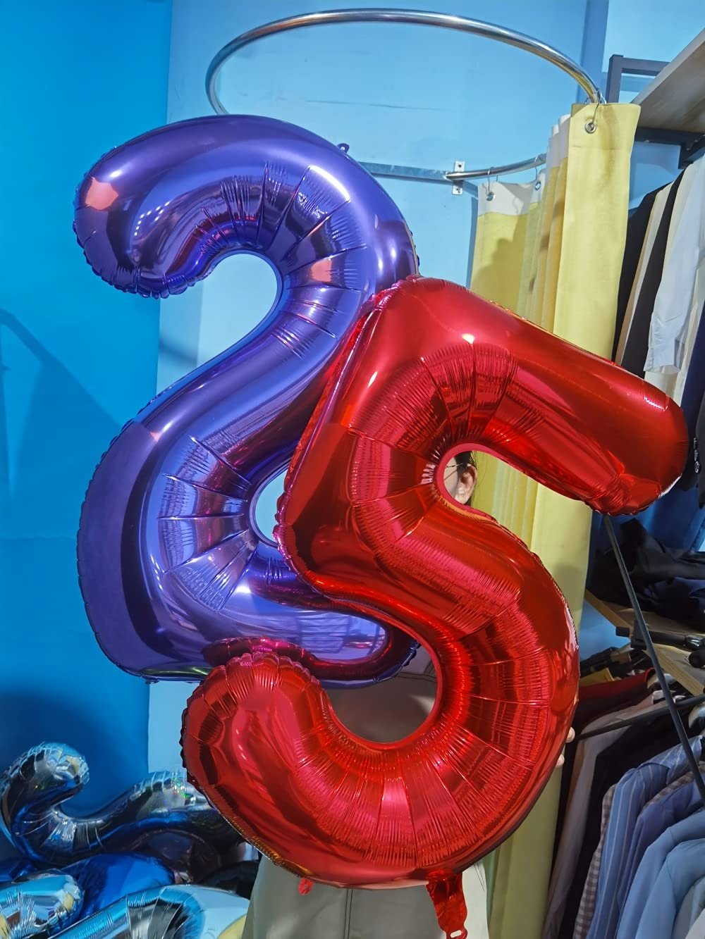 Two large foil number balloons, one red '5' and one purple '2', held up against a blue wall with clothing racks in the background.