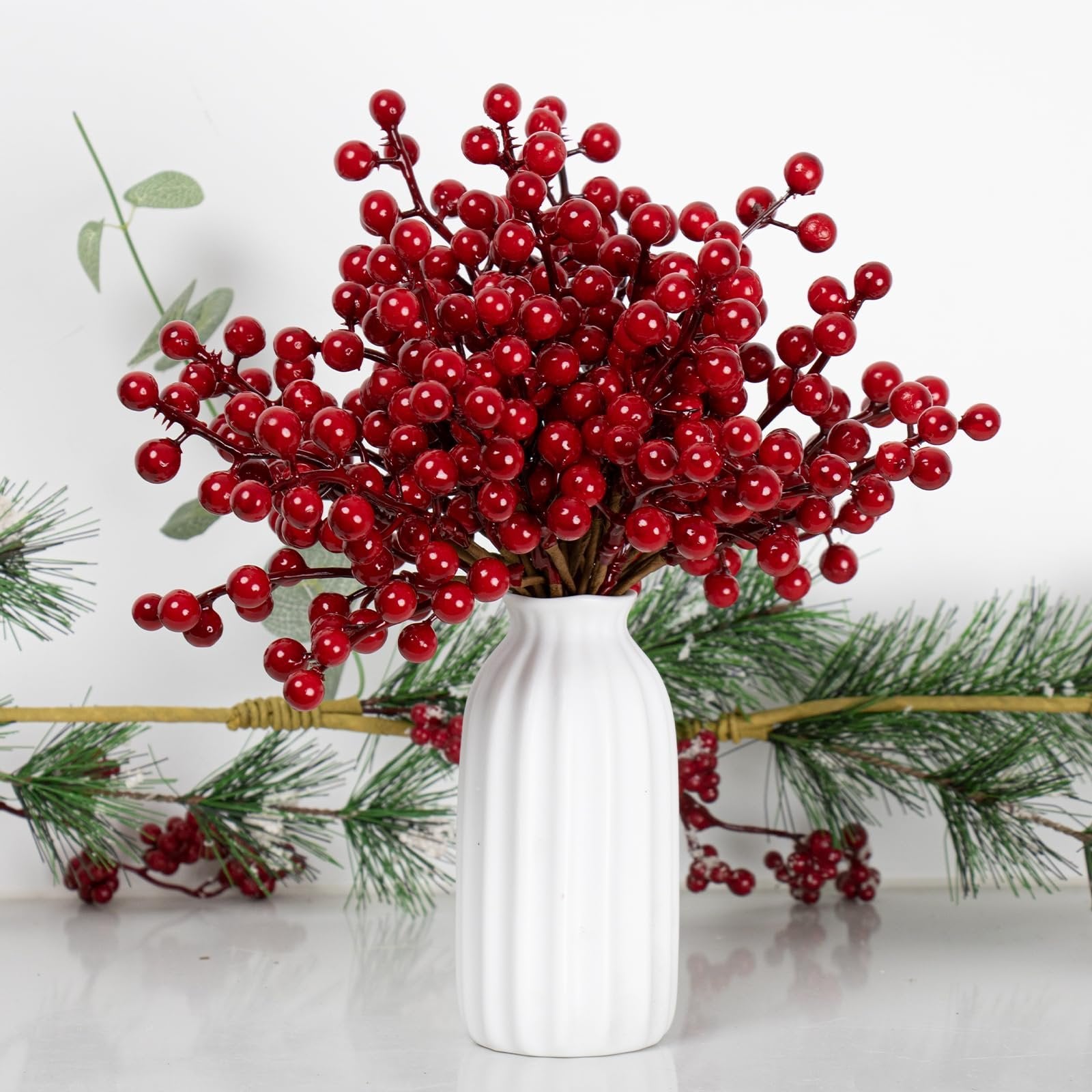 Artificial red berry stems with holly berries in a white vase, with pine branches behind.