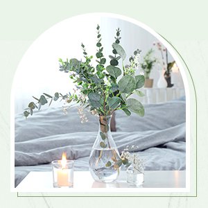 Artificial eucalyptus stems in a clear glass vase with a lit candle and small floral arrangement on a bed.