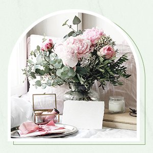 Arrangement of pink peonies and eucalyptus stems in a glass vase on a tray with a jewelry box and candle.