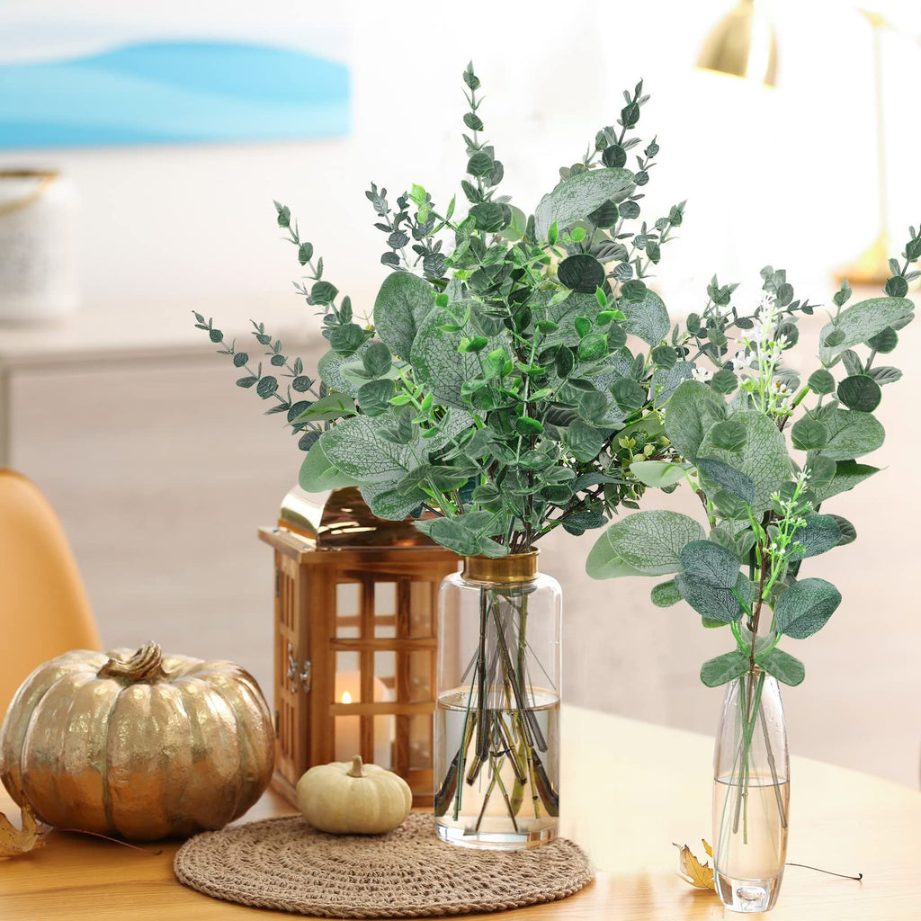 Two vases of artificial eucalyptus stems with silver dollar leaves on a table with decorative pumpkins and a lantern.