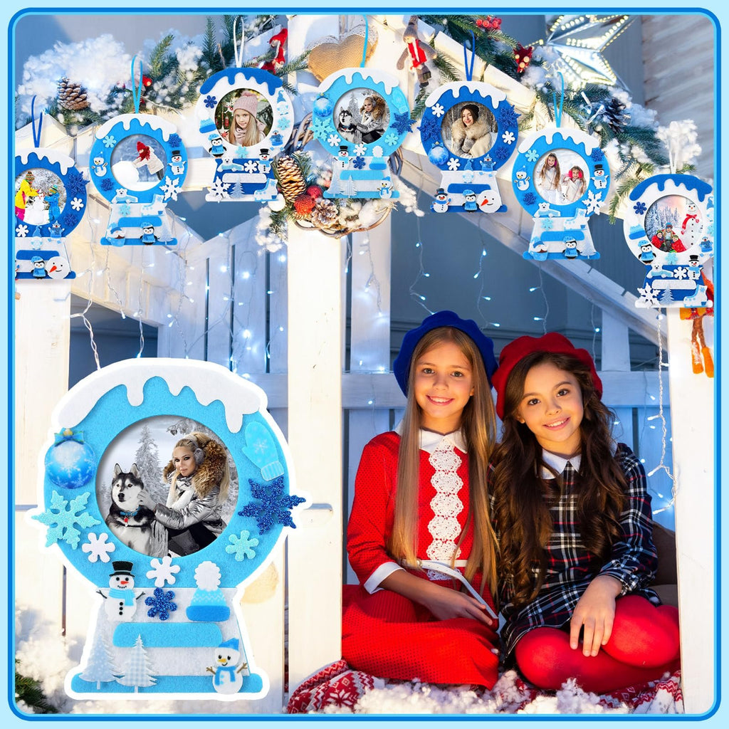 Two girls smiling in front of a display of winter-themed felt picture frame crafts hanging on a railing with fairy lights.