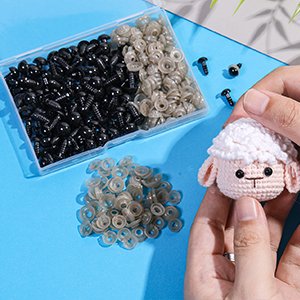 Hands holding a crochet sheep head with black plastic safety eyes and washers scattered around a clear plastic box of eyes.
