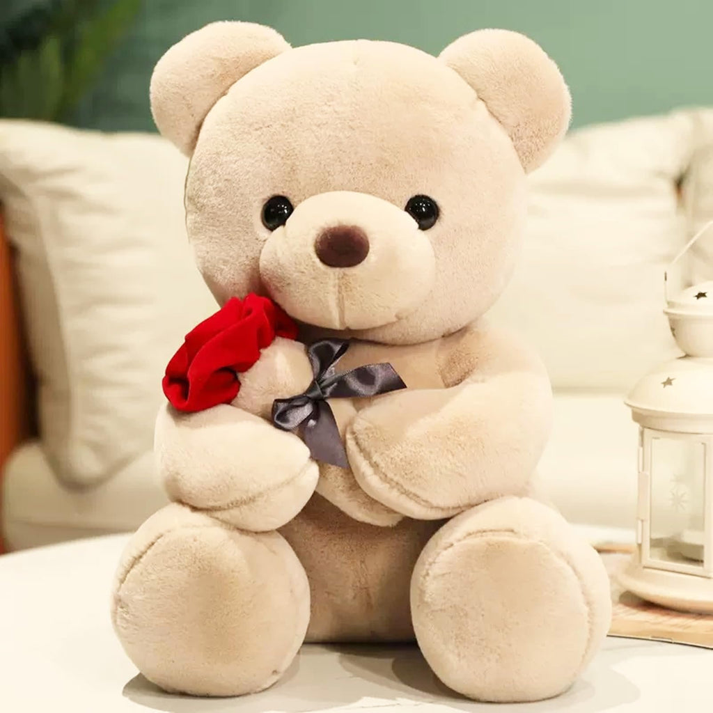 Plush teddy bear holding a red rose and wearing a gray ribbon bow, sitting on a table with a lantern.