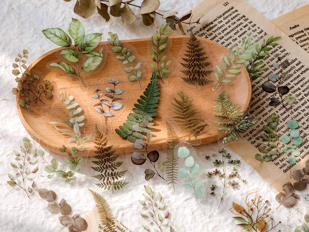 Assorted clear PET botanical stickers of leaves and ferns arranged on a wooden dish and scattered on textured paper.