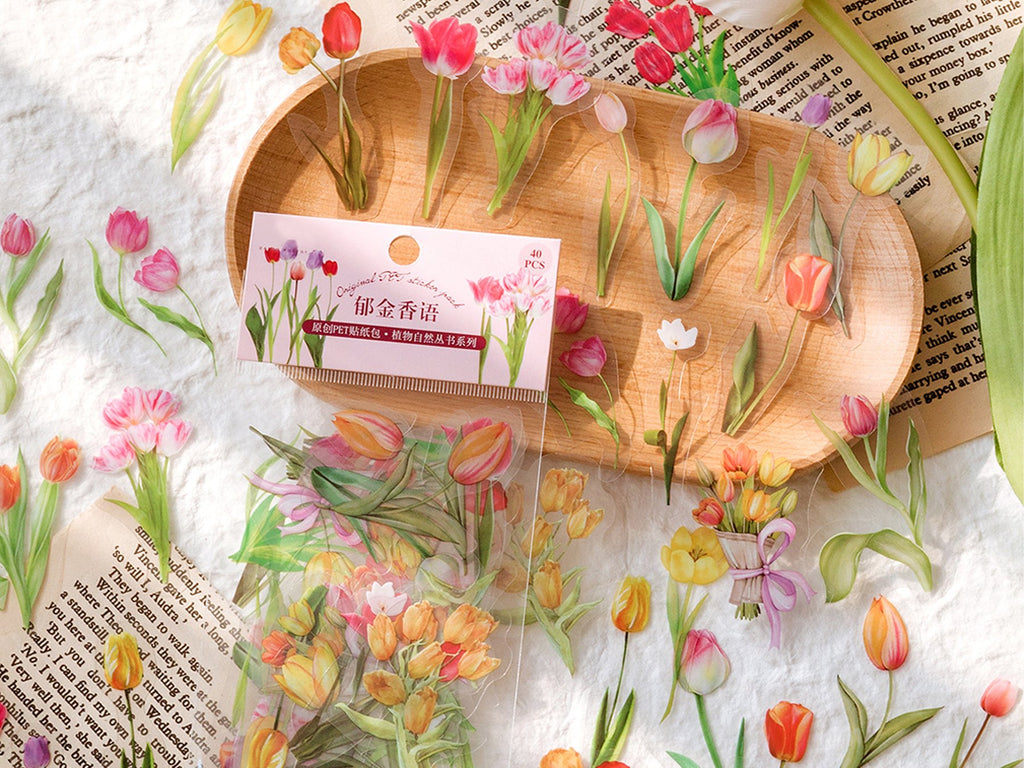 Assorted clear PET botanical stickers of tulips and leaves scattered on a textured surface and a wooden tray.