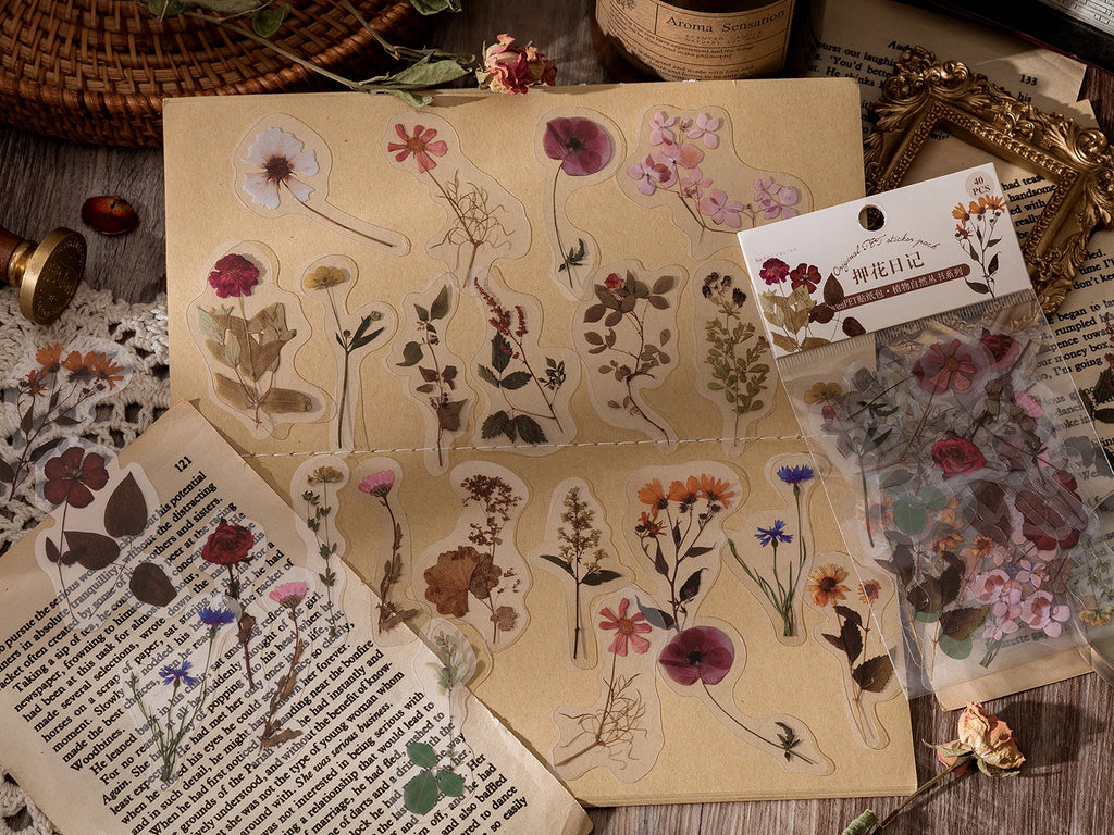 Assorted clear PET botanical stickers of flowers and leaves arranged on a brown paper sheet, with a sticker pack visible.