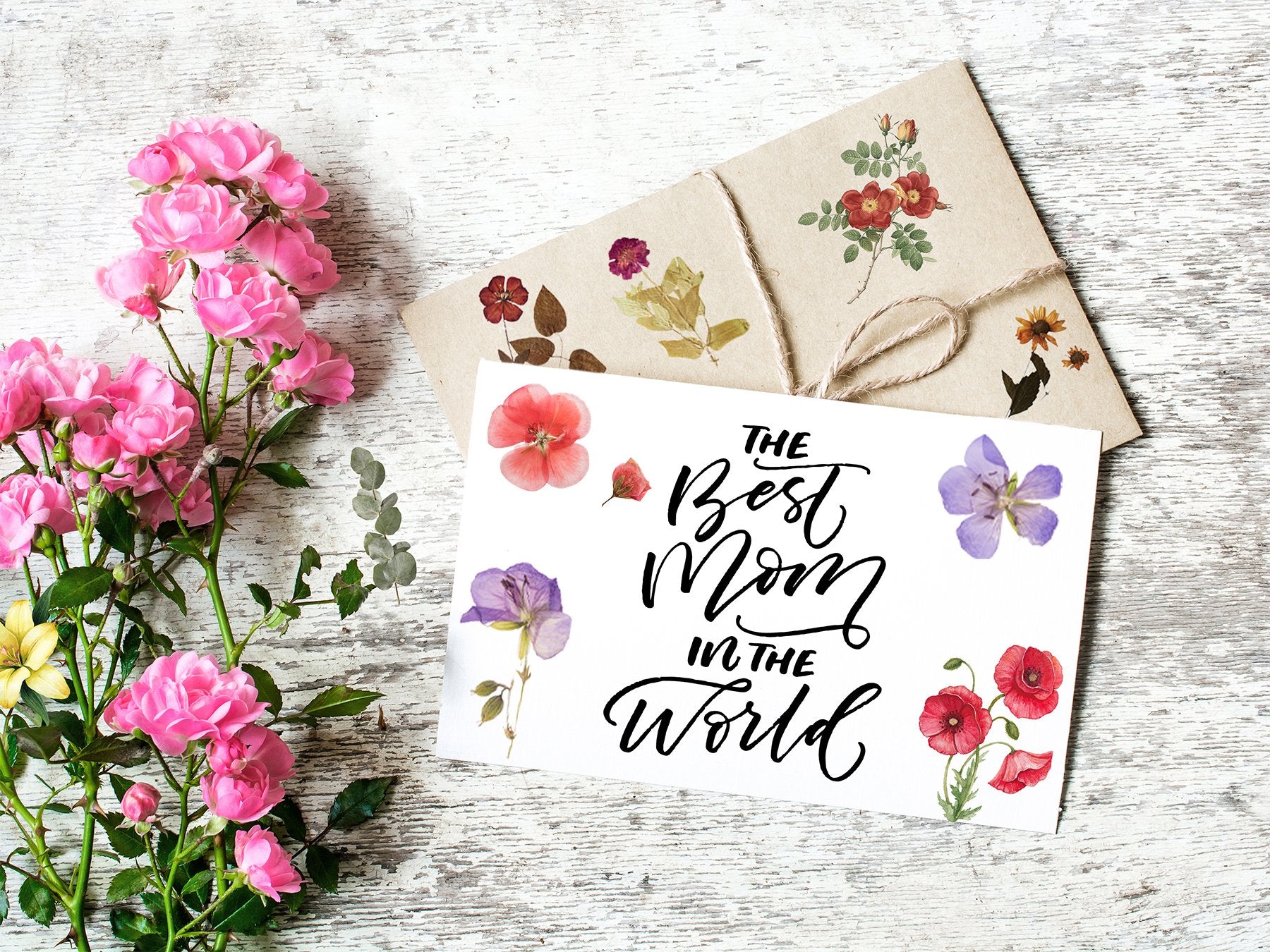 Card with floral stickers and text 'The Best Mom in the World' next to pink roses on a wooden surface