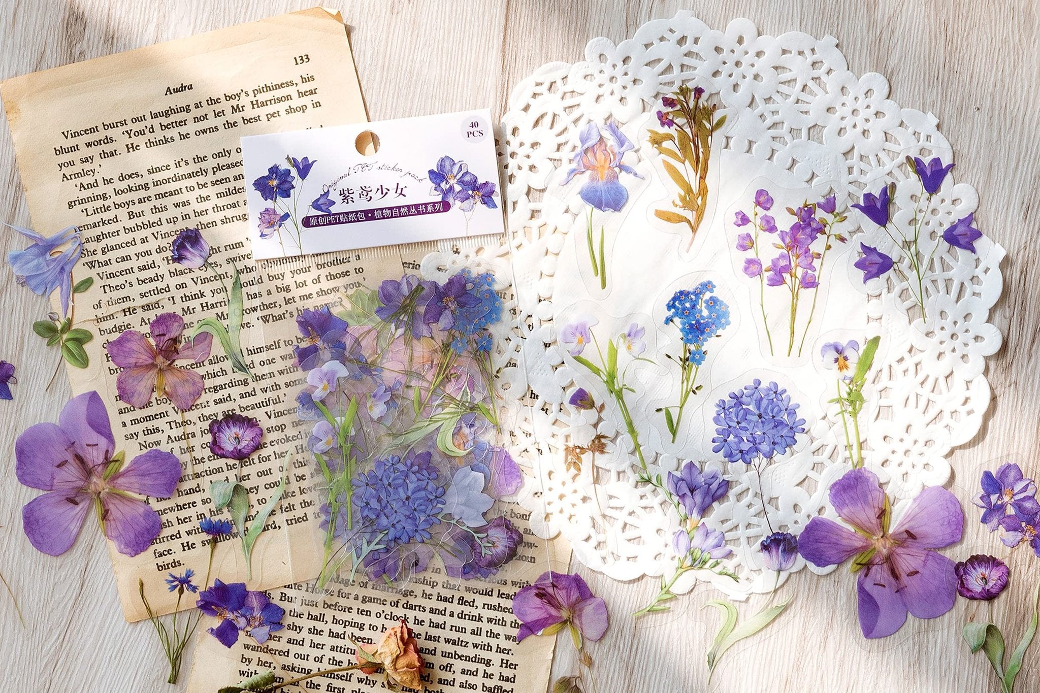 Assorted clear PET botanical stickers of flowers and leaves scattered on a book page and white doily.