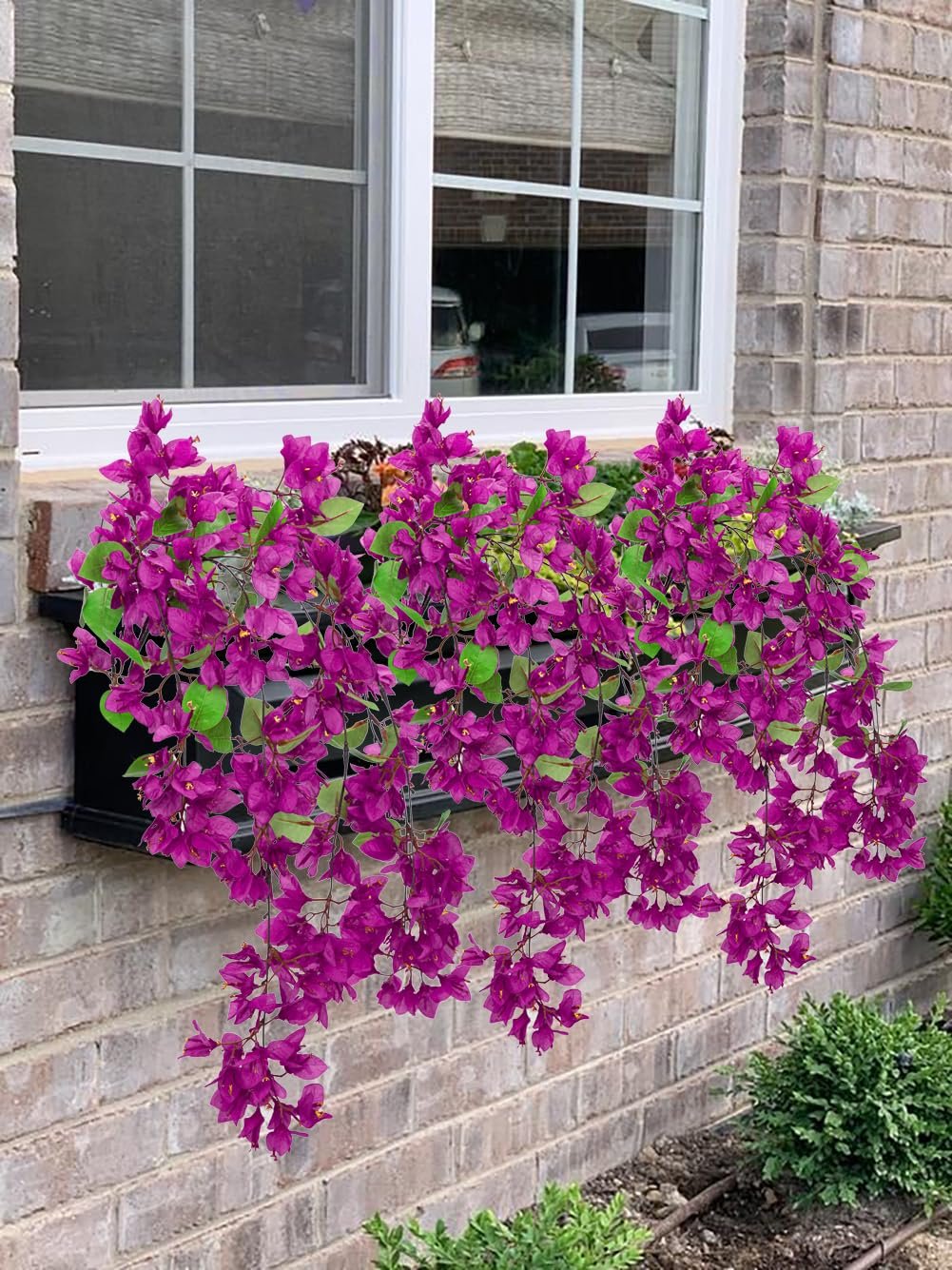 Purple artificial bougainvillea flowers cascading from a window box on a brick wall.