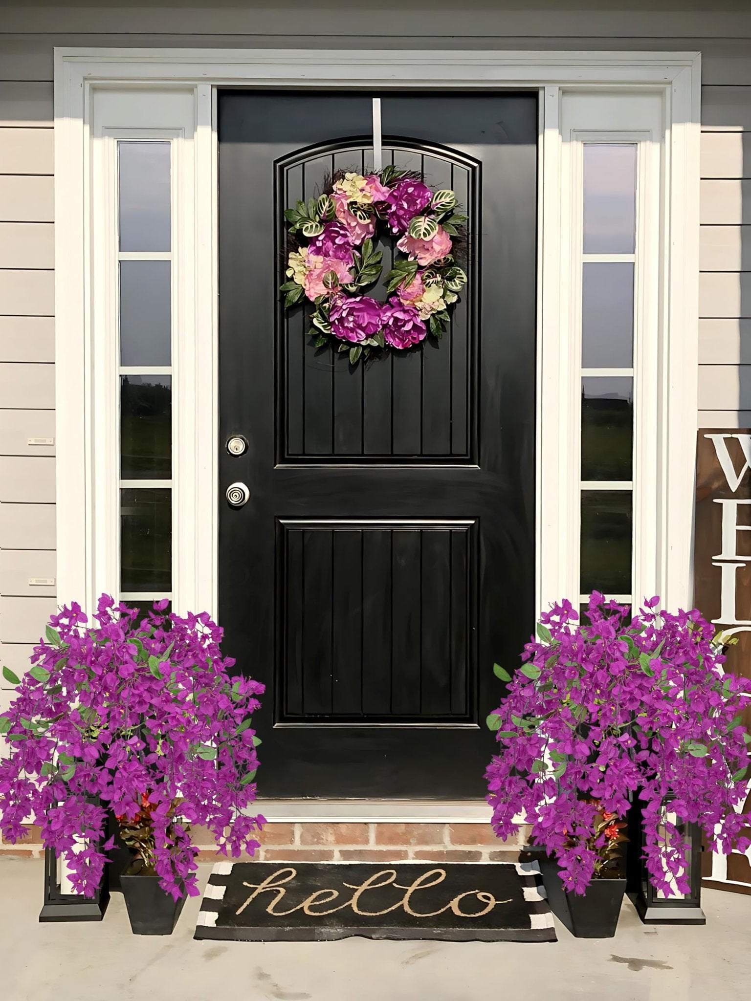 Front door decorated with a floral wreath and two potted purple flowers, with a 'hello' doormat.