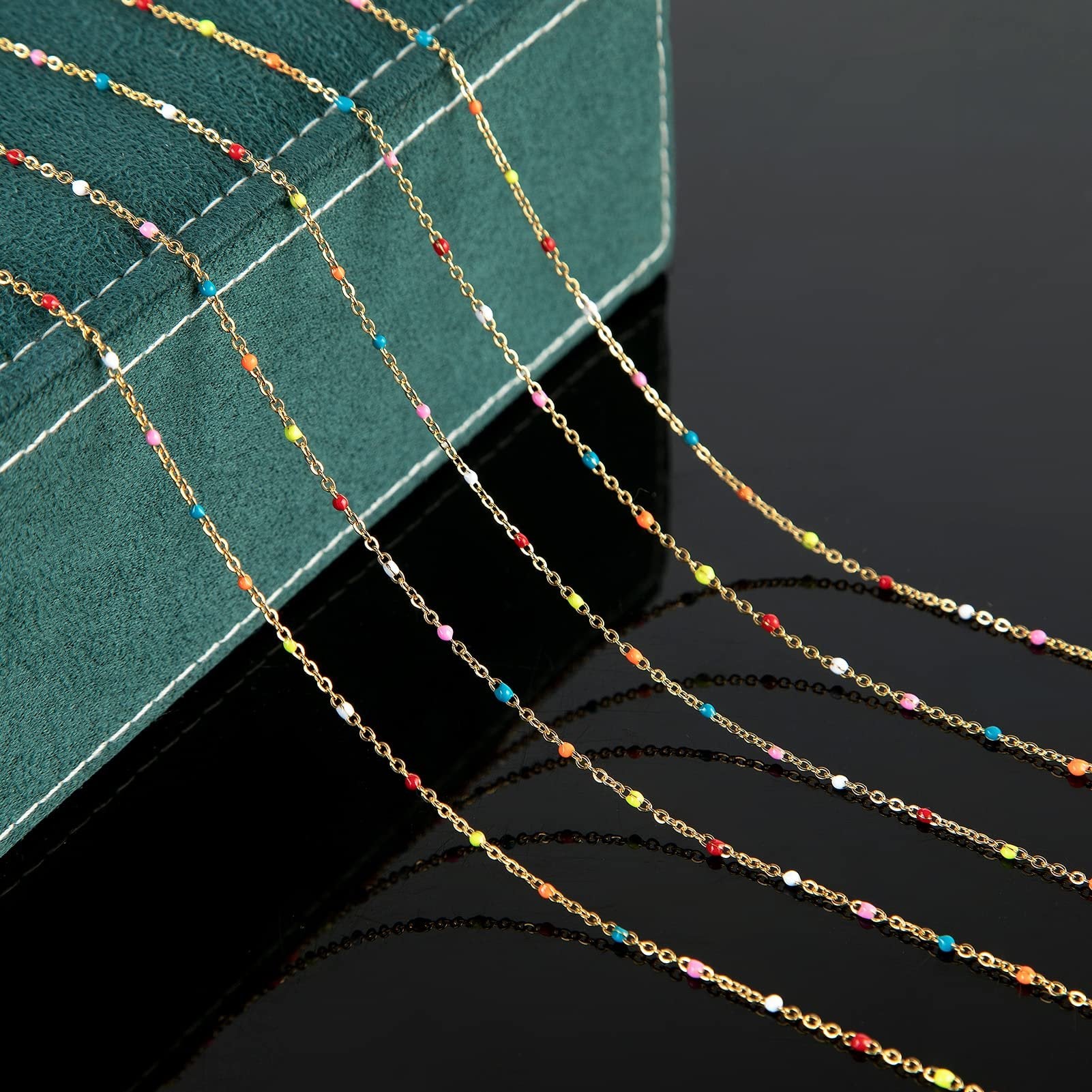 Gold-plated stainless steel beaded jewelry chain with colorful beads, laid out diagonally on a dark surface with a green textured box.